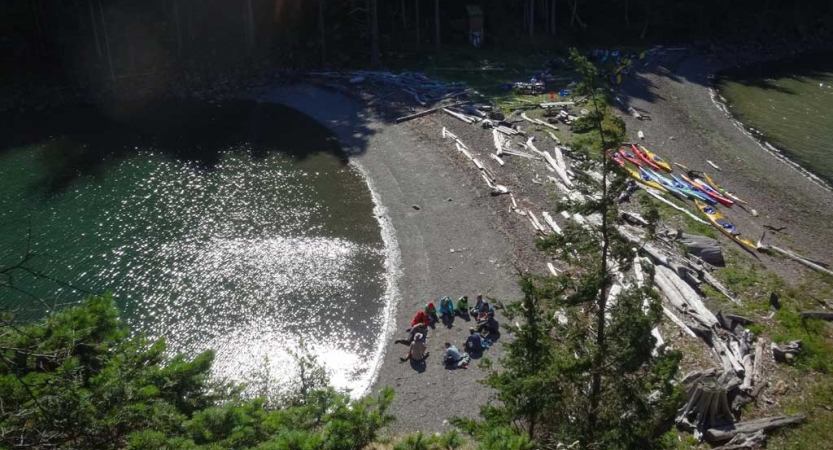 From an aerial point of view, a group of people sit in a circle on a beach. Kayaks and driftwood are also beached nearby.